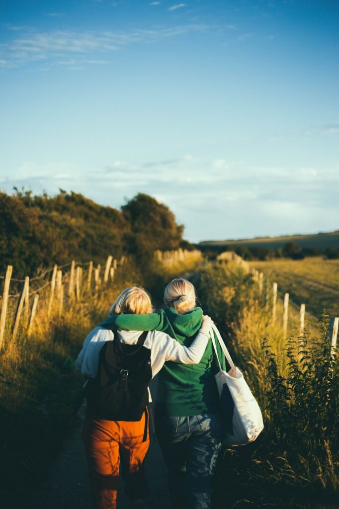 two women with their arms around each other's shoulders walking on a path through a field