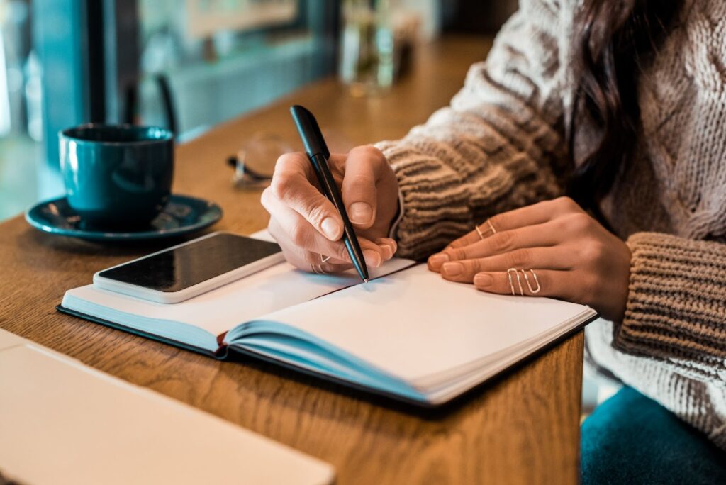 woman sitting at a table in a coffee shop writing in a journal. cell phone and cup and saucer next to her