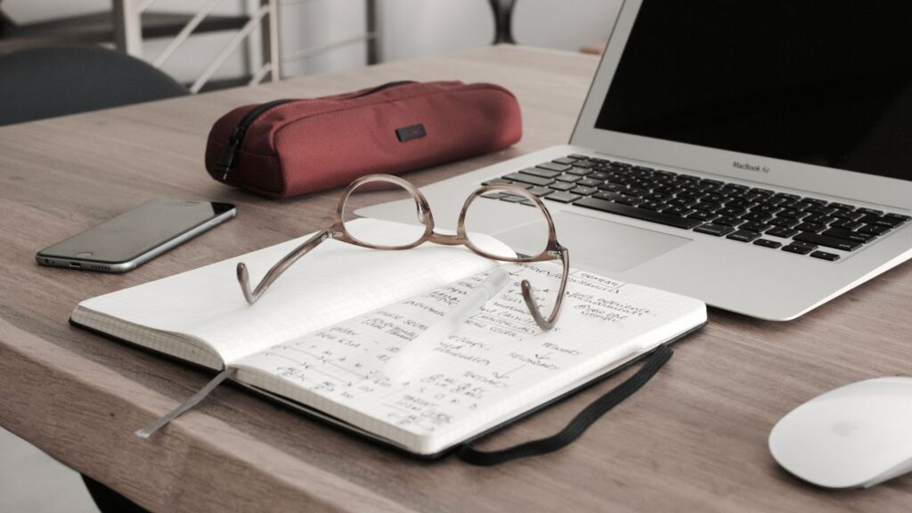 laptop computer on a table. Open journal with eyeglasses on it in front of the computer. Pencil case, cell phone, mouse next to computer.