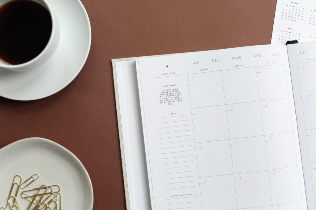 planner open to a monthly calendar page. Cup of coffee on a saucer and small plate holding paper clips on the table next to the planner