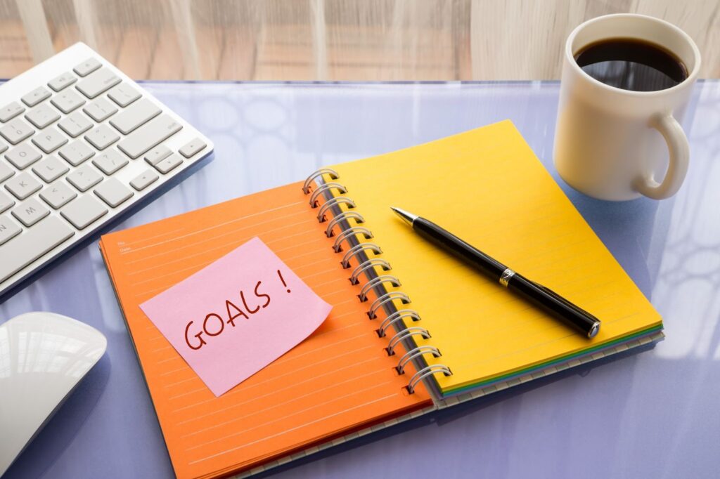 open spiral notebook with orange and yellow pages. Pen and pink post-it note with "Goals!" written on it sitting on the pages. Cup of coffee, computer keyboard, and mouse next to notebook