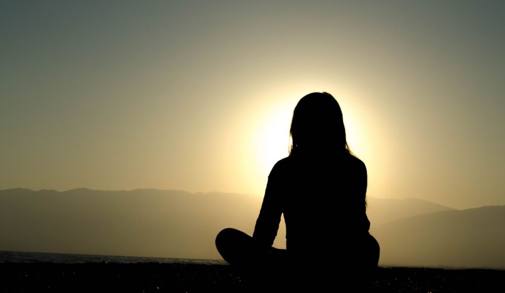 silhouette of a woman sitting cross-legged on the ground at sunrise or sunset. mountains in the background