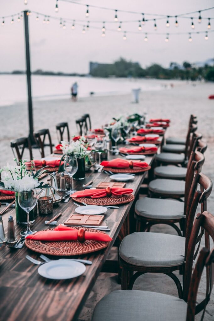 long dining table on the sand near a body of water. Table is set with red placemats & napkins, gray bread plates, and wine glasses