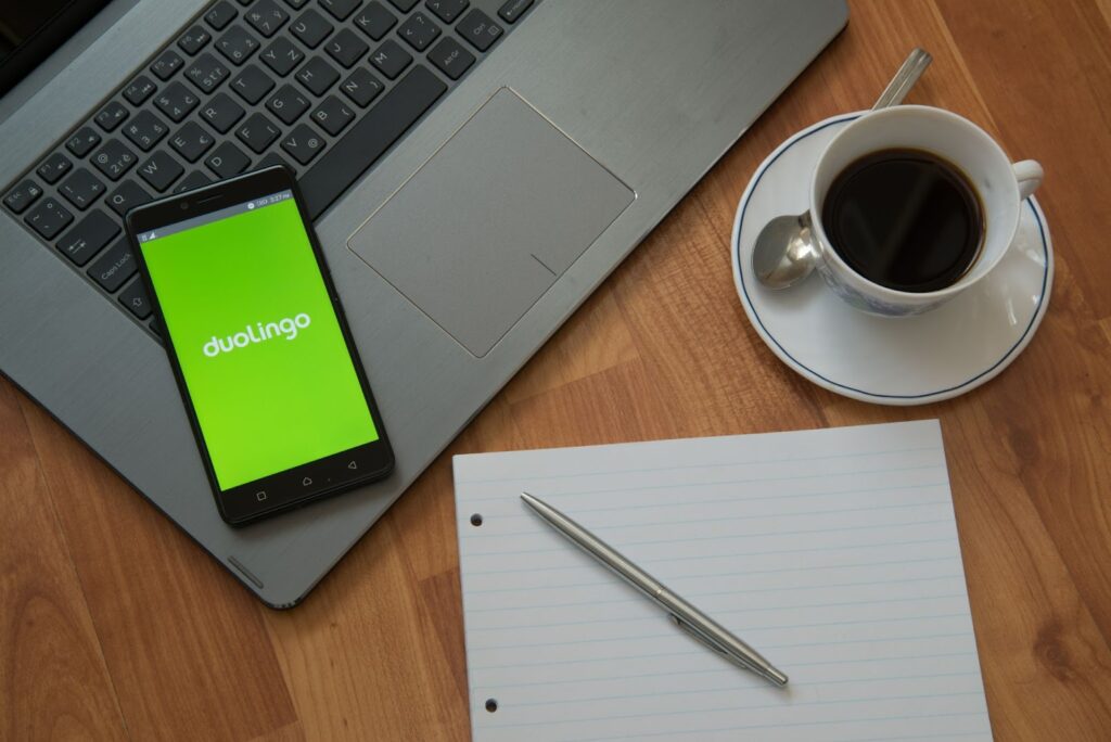 keyboard portion of a laptop computer with a cell phone resting on it. Phone screen shows logo for language learning app DuoLingo on a bright green background. Cup of coffee and spoon on a saucer, and notebook paper and a pen are next to the computer