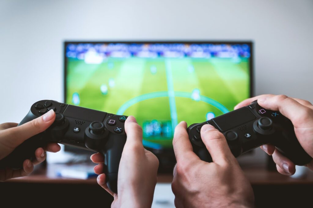 two hands holding video game controllers in the foreground; tv showing soccer video game in the background