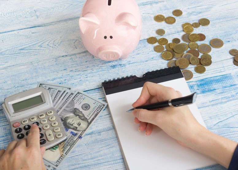 right hand holding a pen about to write on a notepad. left hand using a calculator. top of image: piggy bank and gold coins