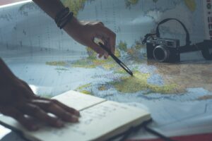 map on table. hand holding pencil pointing to map; other hand holding an open journal; camera in the background