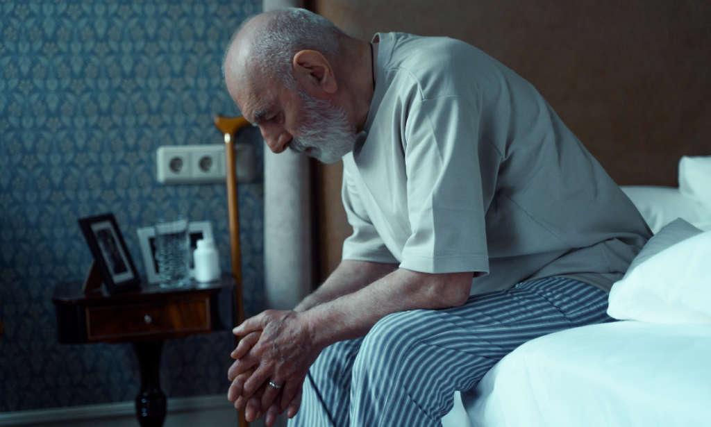 older man sitting hunched over on the side of a bed, looking sad or bored