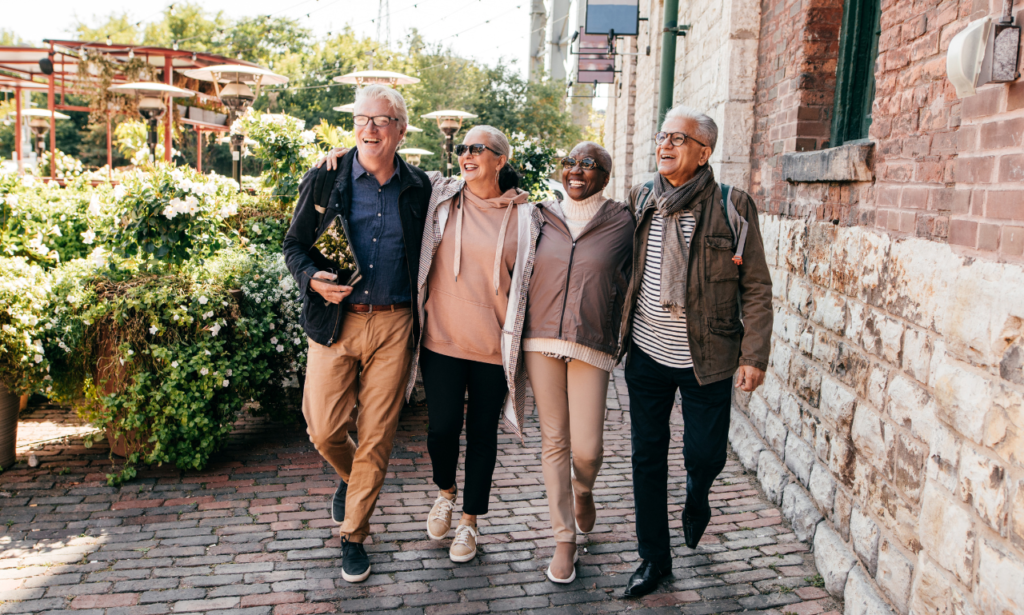 group of four older people walking with arms around each others' shoulders. On a stone path next to a stone building with greenery and tall pathway lights on the left