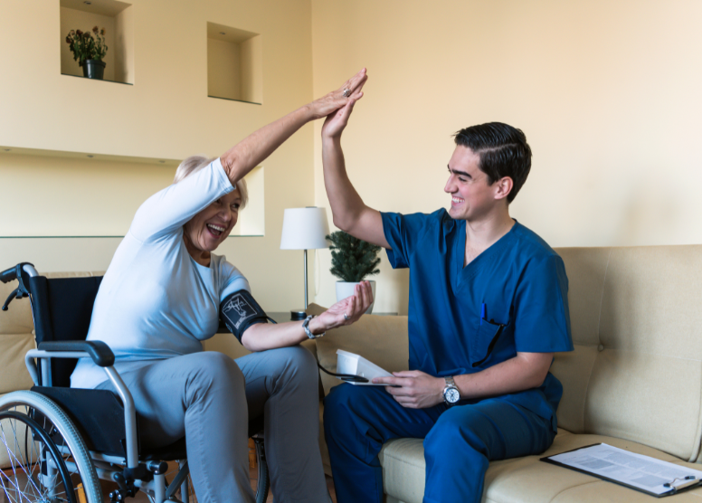 woman in wheelchair giving a high five to a male caregiver who is sitting on a couch next to her taking her blood pressure