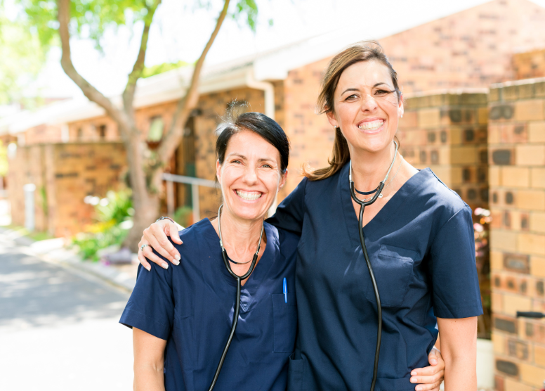 two female caregivers wearing scrubs and with stethoscopes around their necks standing with their arms around each other