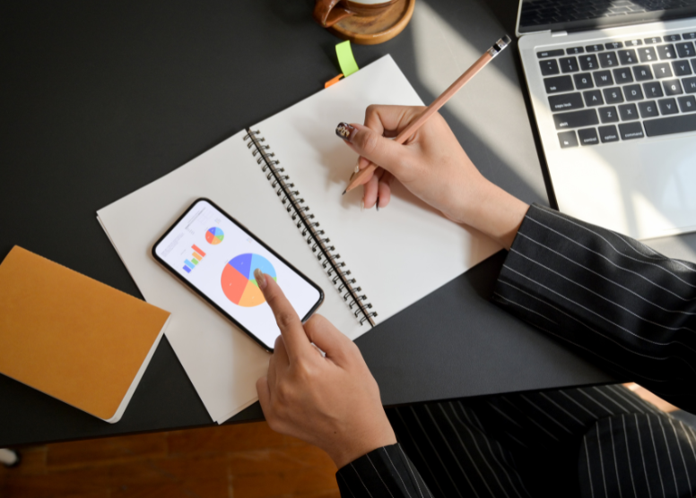 overhead view of a desk with a woman sitting at it (only hands, forearms visible). woman holding a pencil poised over a spiral notebook page and pointing to a pie chart on a cell phone with her other hand. laptop computer and a small notebook also on table