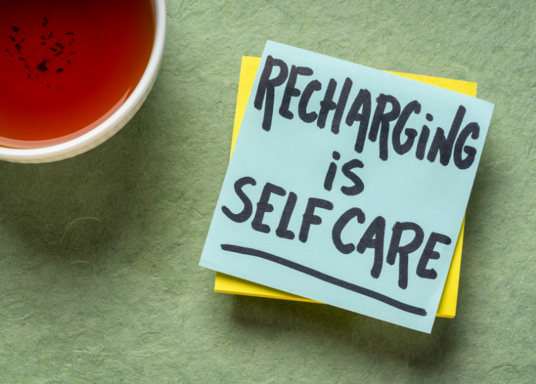 cup of tea in upper left corner of image; post-it in center with words "Recharging is Self Care" written on it