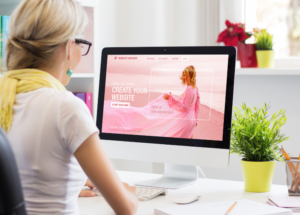 woman sitting at a desk looking at a computer monitor showing a woman in a pink dress on a pink beach and the words "create your website"