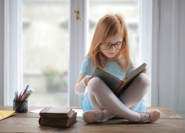 little girl in a blue dress and ballet slippers with red hair and glasses sitting cross-legged on a table reading a book. Two books stacked next to her, and a pencil cup filled with pencils and markers