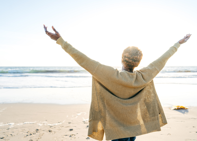 woman standing on the beach facing the water, arms raised up