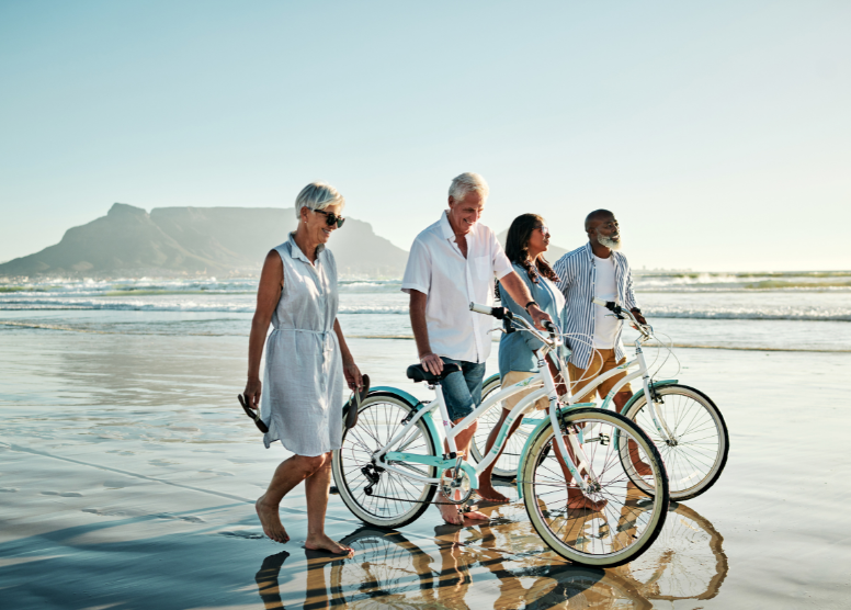 2 couples walking on the beach. Each man is walking a bicycle.