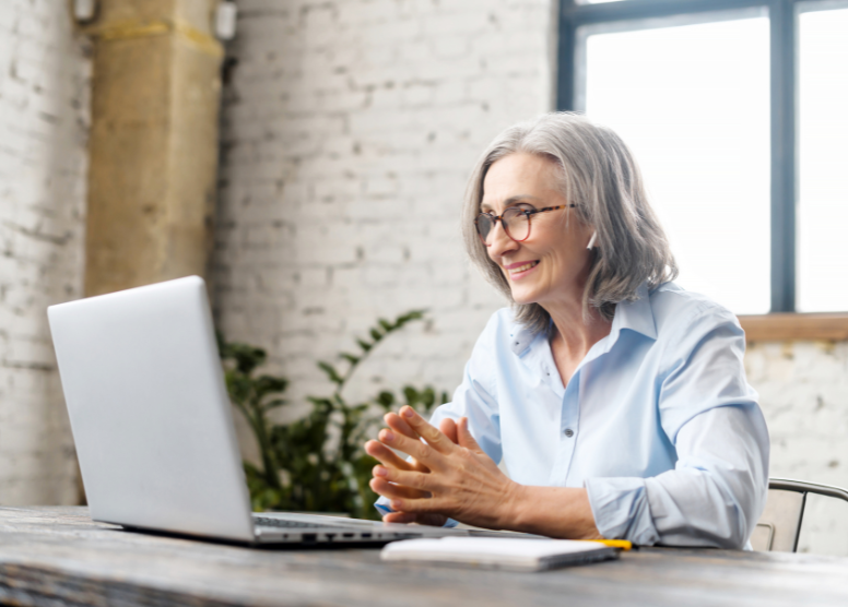 woman with gray hair and glasses sitting at a table in front of a laptop. Wearing earbuds and smiling at the screen like she's talking to someone