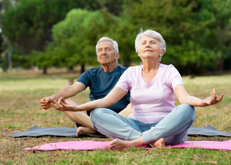 older man and woman sitting cross-legged on yoga mats outdoors, eyes closed, arms resting on knees (meditating)