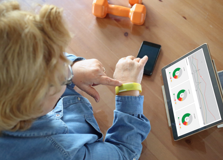 woman touching a yellow fitness tracking on her wrist. Tablet computer open on the table showing graphs and pie charts. Cell phone and orange hand weight on table.