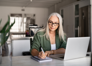 Woman with long silver hair and glasses sitting at a table in front of a laptop computer and writing in a spiral notebook