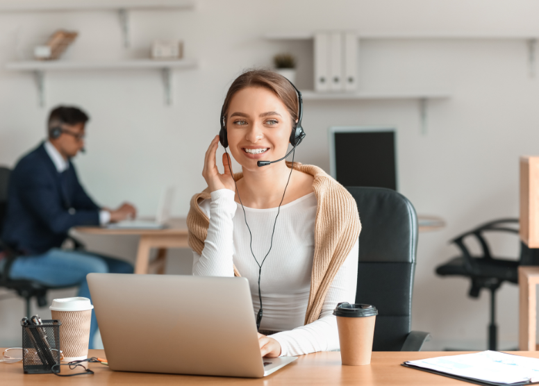 Woman in a customer support office sitting in front of a laptop and wearing a headset with a microphone. Cup of coffee in a to-go cup on the table