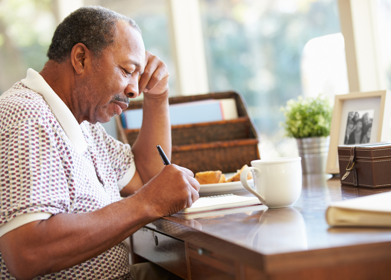 older man sitting at a desk writing in a spiral notebook with a pen. Things around him on the desk: coffee cup, book, leather box with a buckle, family photo, plant, basket with papers in it, plate with food on it