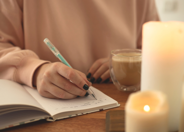 woman writing in a journal with a pen. two candles in the foreground. clear glass mug of coffee next to her