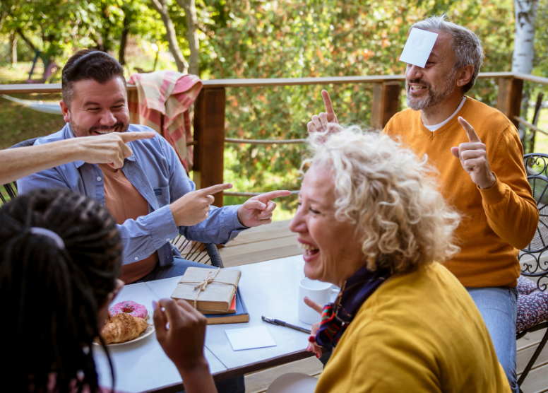 group of people sitting at a table on a porch, playing a game. One man has a yellow post-it on his head; other people are pointing to him. Everyone is laughing