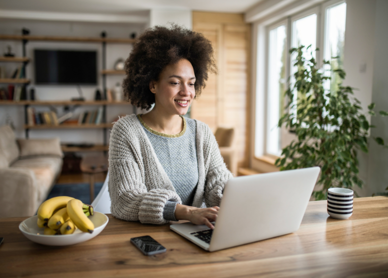 woman sitting at a table typing on a laptop computer. Striped mug is to her left, bowl of bananas and cell phone to her right. Background is a living room area.