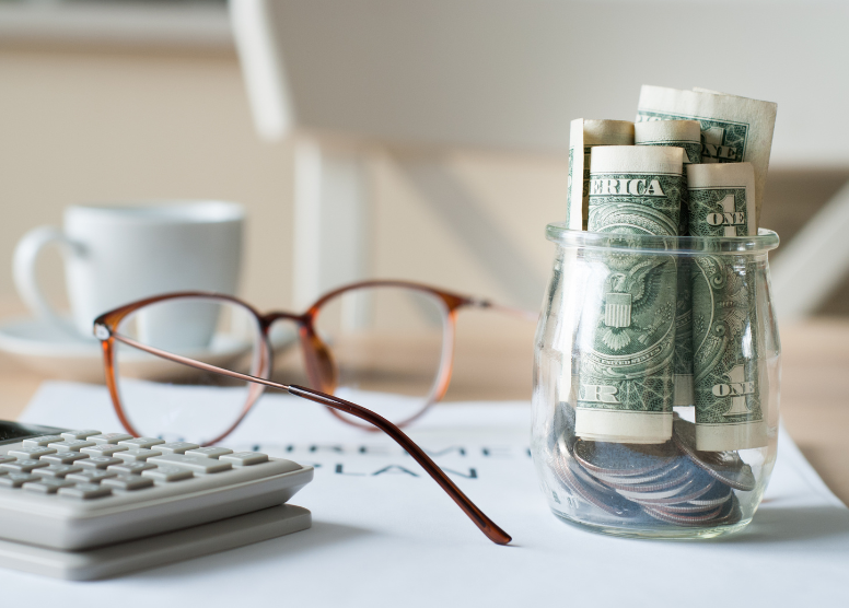 small glass jar with rolled up dollar bills and change in it, sitting next to a pair of glasses and a calculator on a desk. Coffee cup and saucer in the background