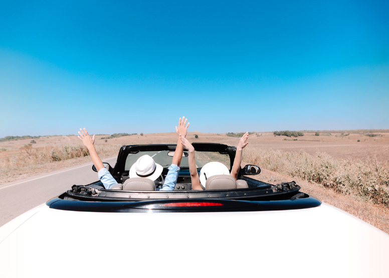 shot from behind of two people wearing hats in a white convertible with the top down. They have their hands in the air. Car is on a highway in the desert with blue sky above