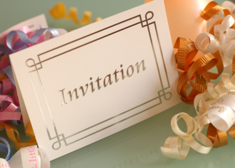 party invitation standing up on a seafoam green surface. Word "Invitation" in gold on front of card. Curled ribbon (orange, white, purple, pink) scattered around card