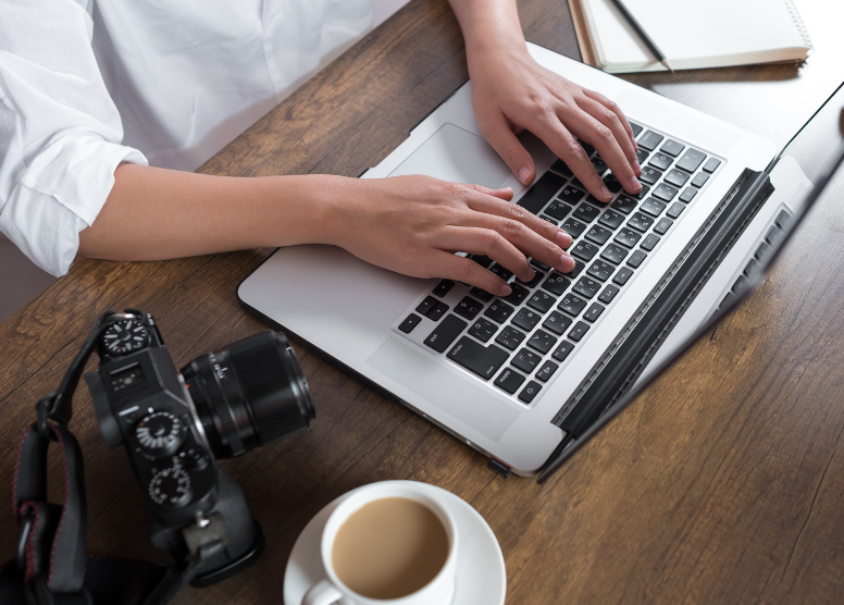 top-down view of woman typing on a laptop. Digital camera, cup of coffee, and notepad with pencil sitting on table surrounding the laptop