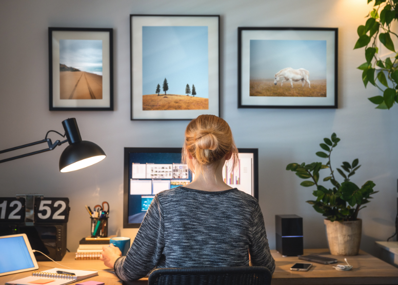 rear view of a woman sitting at a desk in front of a large computer monitor surrounded by a lamp, a plant, and various office supplies. 3 framed pictures on the wall