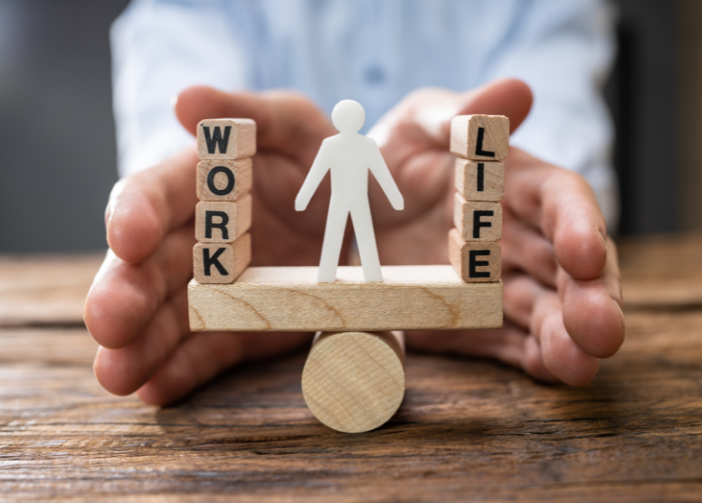 small wood letter cubes stacked to form the words "WORK" on the left and "LIFE" on the right, balanced on a rectangular wood block that is balanced on a wood cylinder. White figure of a man in between them