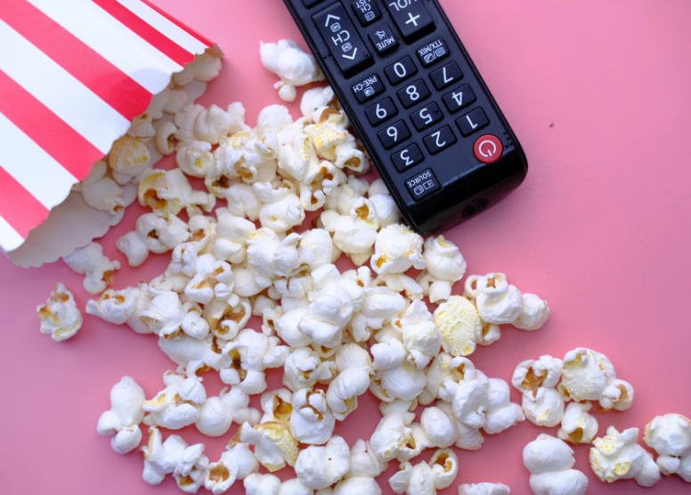 remote control next to popcorn spilled out of a red and white striped bag. Pink background