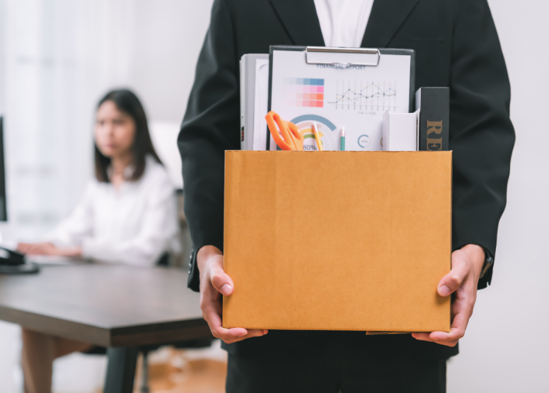 man (from the chest down) holding a box of desk contents. woman sitting at a computer out of focus in the background