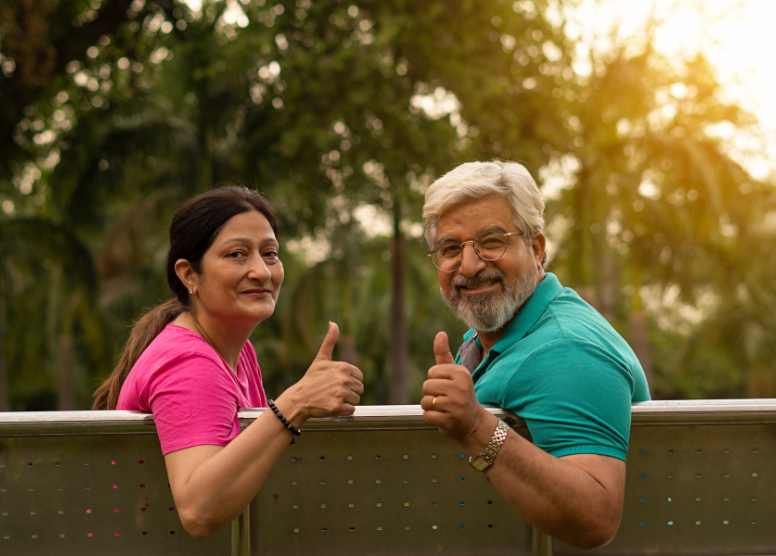 man and woman retirees sitting on an outdoor bench. Turned around facing backward, smiling and giving thumbs up