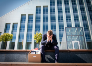 man sitting on steps outside a tall building with his head in his hands. box of desk contents sitting next to him