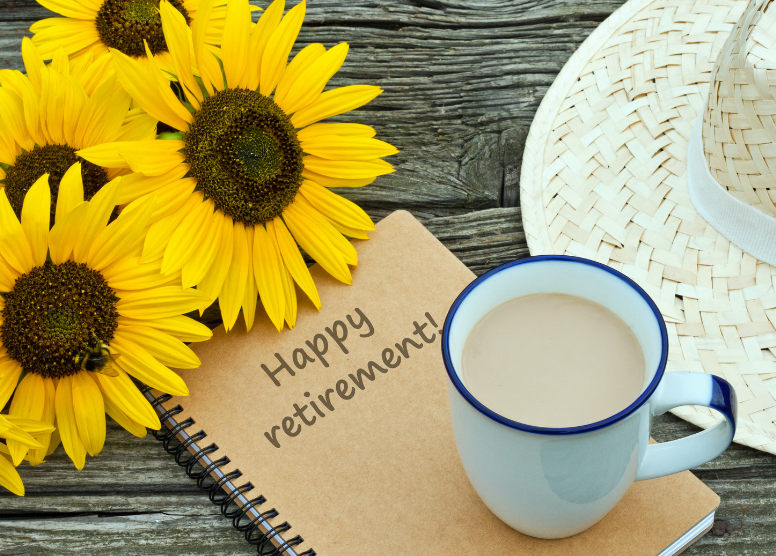 wood plank table with spiral notebook with the words "Happy retirement!" on it; cup of coffee sitting on notebook; straw hat and sunflowers at top and side of notebook