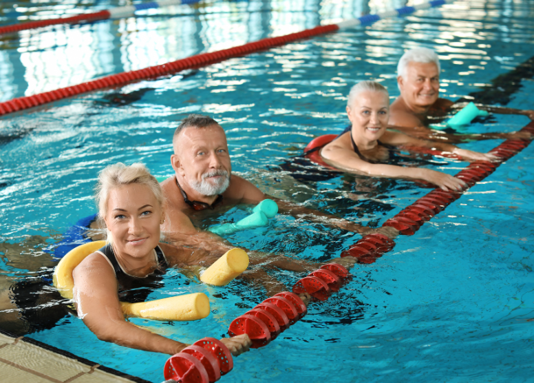 group of 4 older people in an indoor swimming pool, holding on to the lane divider and using pool noodles