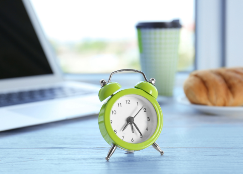 green retro alarm clock sitting on a table. laptop, to-go coffee cup, and bread sitting on a plate are out of focus in the background