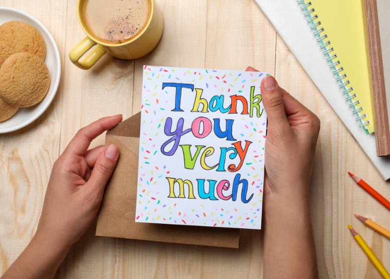 person holding a card that says "Thank you very much". Plate of cookies, mug of coffee, notebooks, colored pencils around on the table