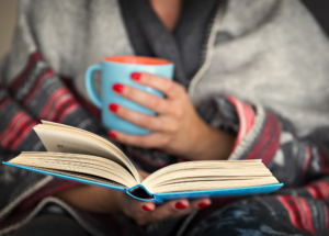 close-up shot of a woman holding an open book in her lap. Holding a blue mug in her other hand